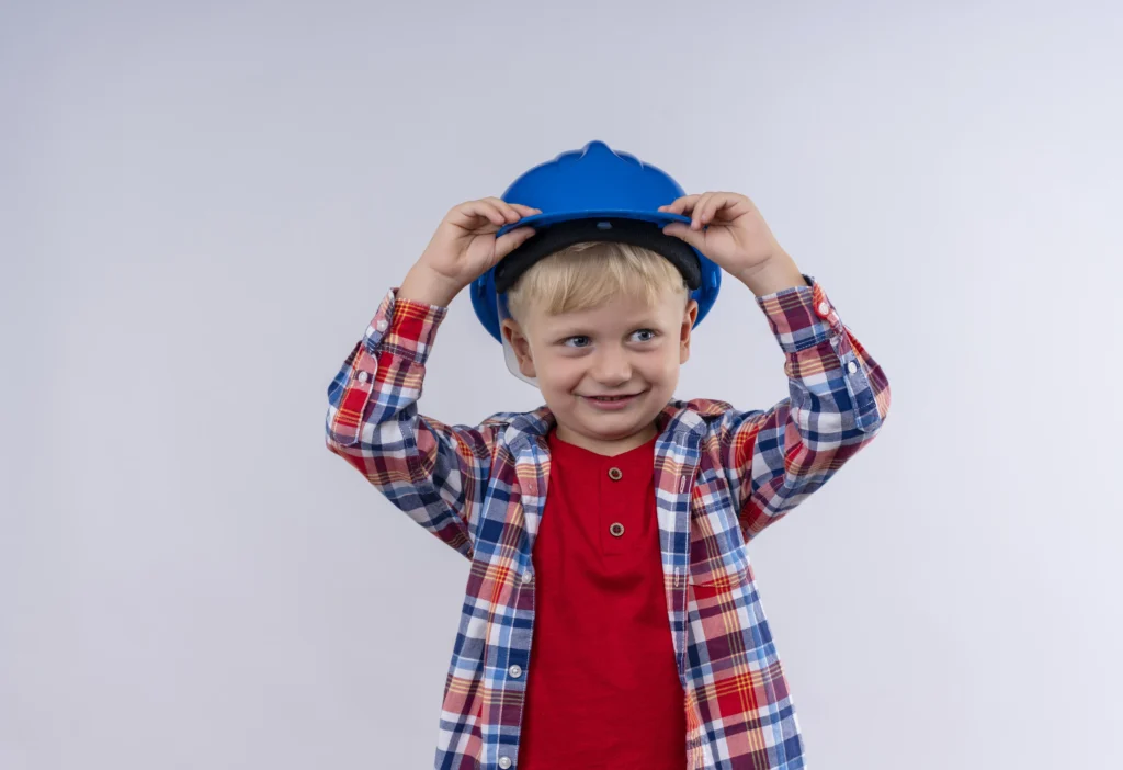 smiling cute little boy with blonde hair wearing checked shirt holding his blue helmet with hands head white wall 1024x702 1